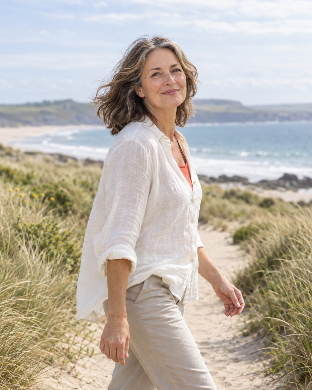 A woman in midlife walking along a bright coastal path, with her hair moving in the breeze